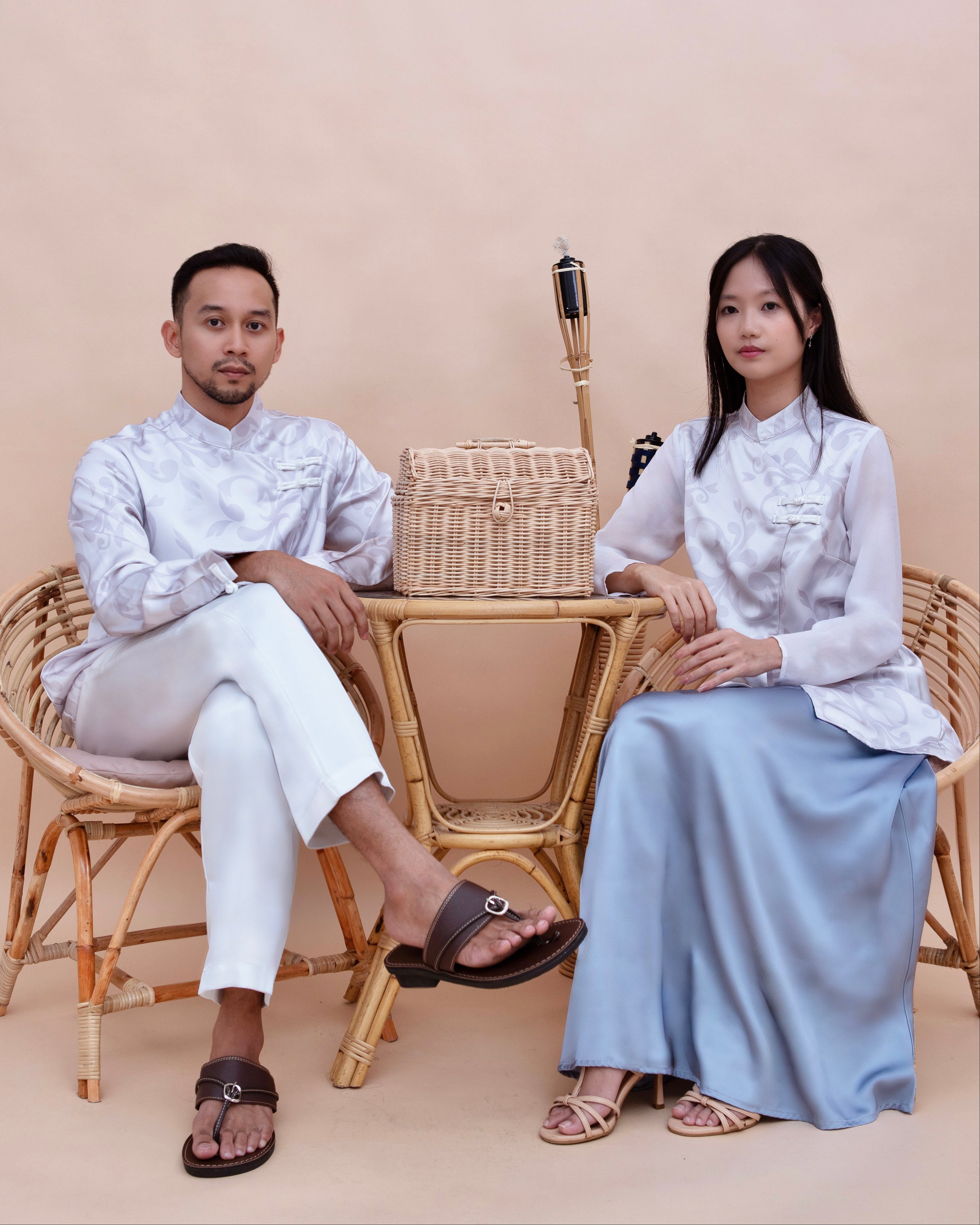 Two people sitting on wicker chairs with a woven basket between them against a beige background.